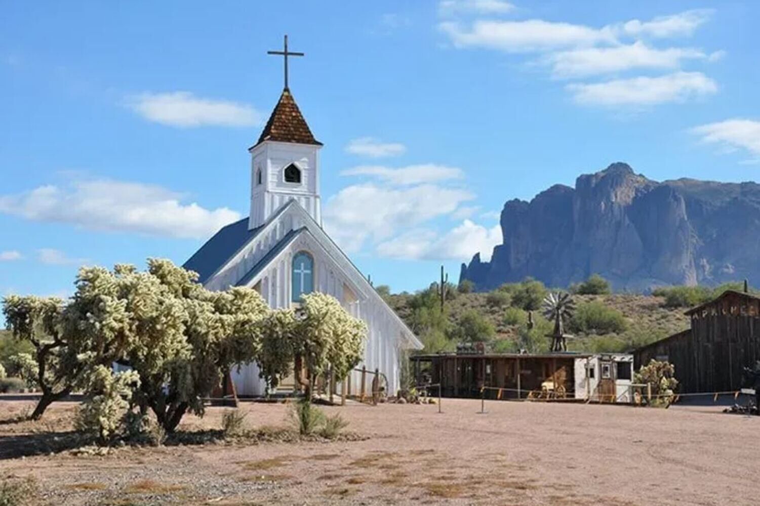 A white church with a cross on top, surrounded by desert vegetation and a mountain in the background.