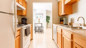A modern kitchen with wooden cabinets, a stove, and a sink. There is a dining area with two chairs and a table. A plant is visible in the background.