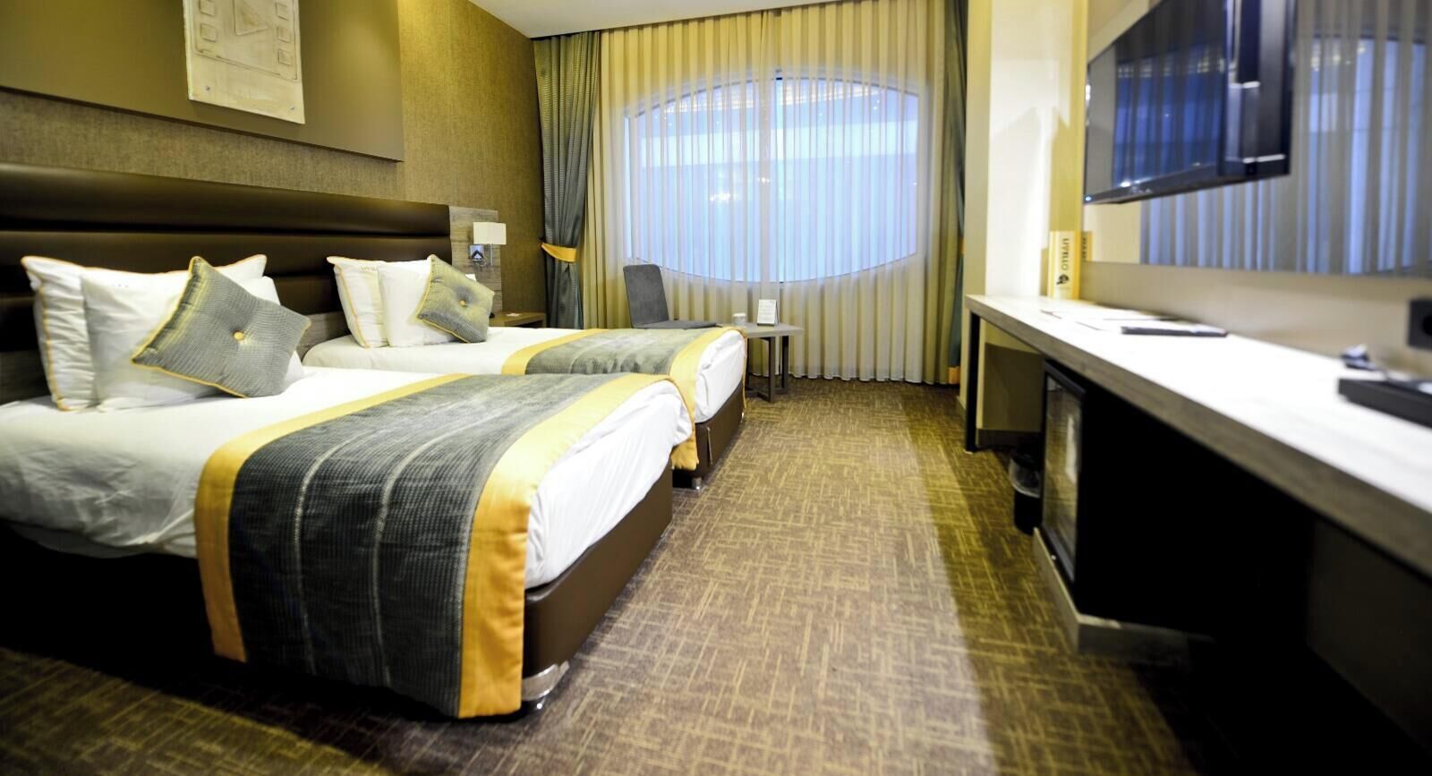 A hotel room with two beds, a desk, a chair, a television, and a large window with curtains.
