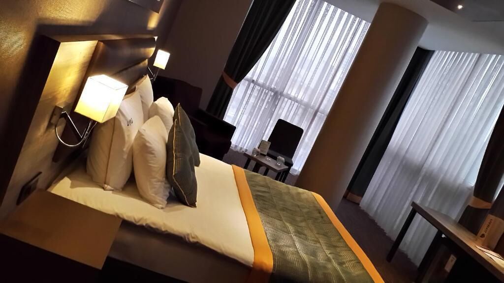 A hotel room with a bed, pillows, a bedside table, a lamp, and a window with curtains.