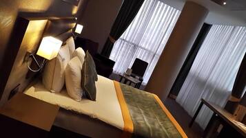 Una habitación de hotel con una cama, almohadas, una mesita de noche, una lámpara y un ventanal con cortinas.