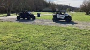 Two off-road vehicles parked on a gravel path in a grassy area with trees and a building in the background.