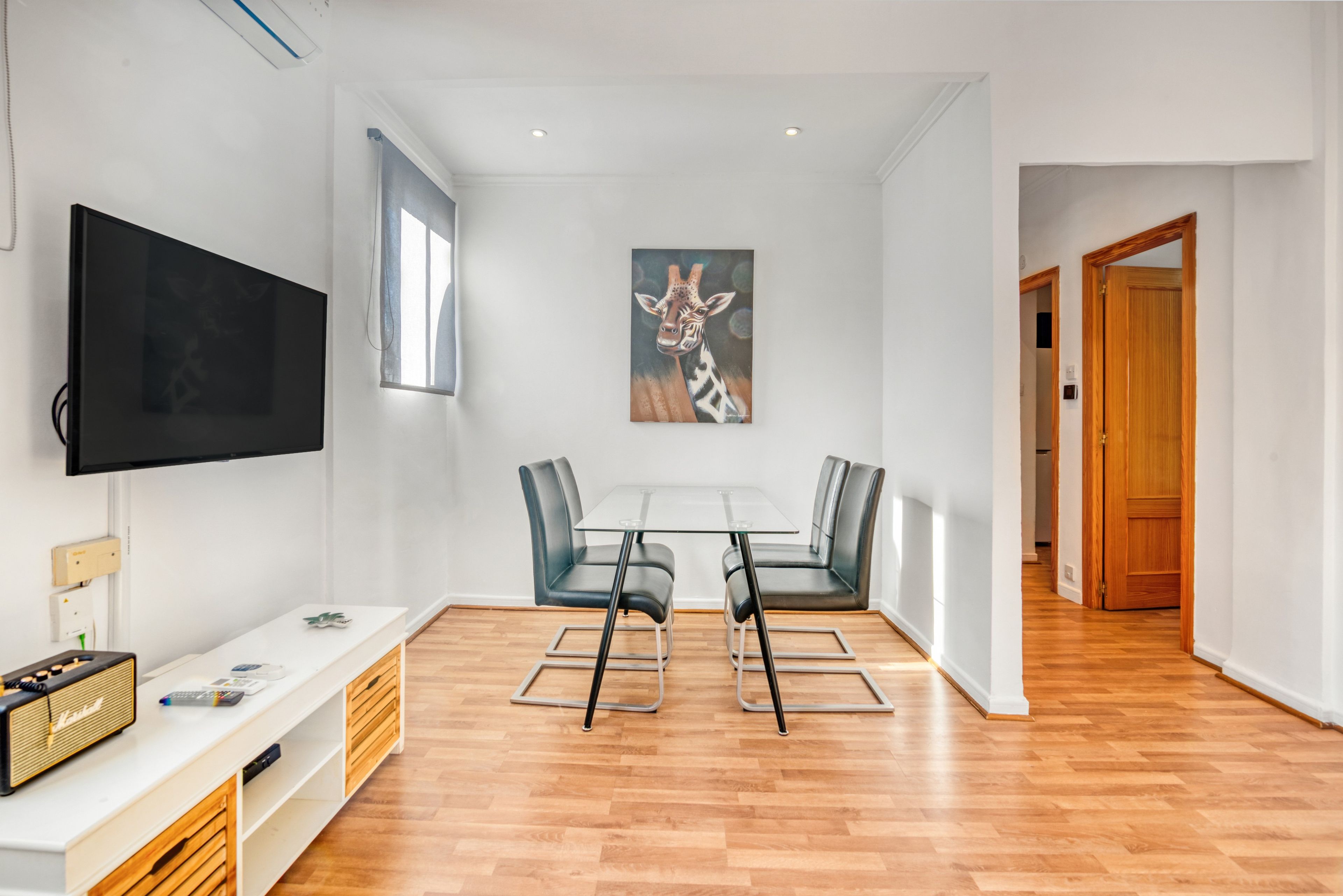 A modern living room with a flat-screen TV, a dining area with a glass table and chairs, and a hallway leading to a wooden door.