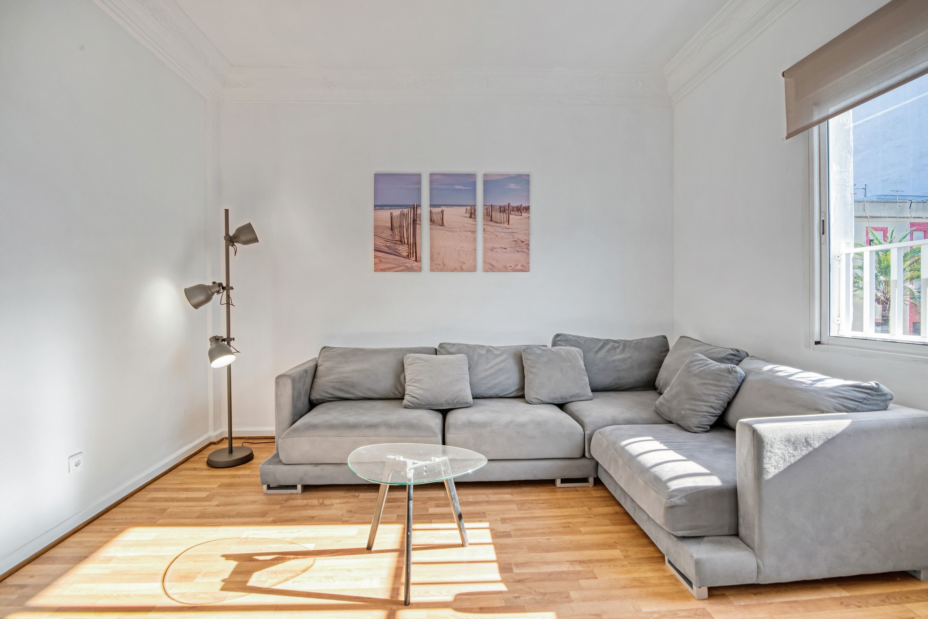 A modern living room with a grey sectional sofa, a glass coffee table, and a floor lamp. There are three framed pictures on the wall.