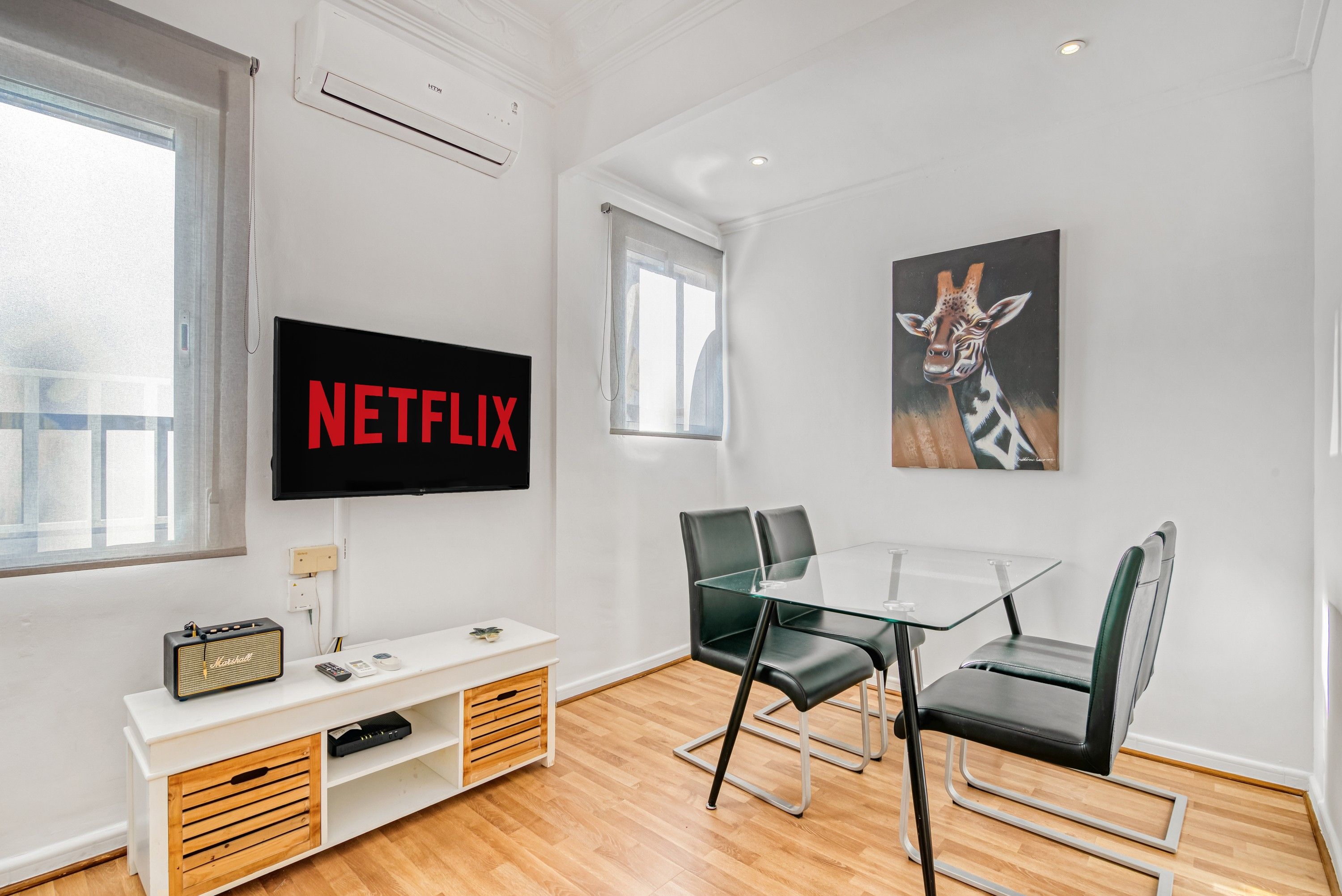 Un salon moderne avec un téléviseur à écran plat fixé au mur, une table en verre avec des chaises, un meuble de rangement blanc avec une radio ancienne, et un tableau représentant une girafe accroché au mur.