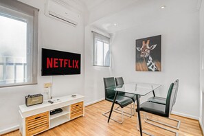 Een moderne woonkamer met een flatscreen televisie aan de muur, een glazen tafel met stoelen, een witte dressoir met een vintage radio, en een giraffen schilderij.