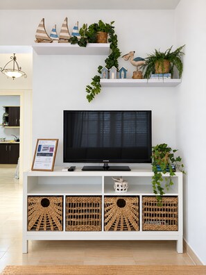 Een moderne woonkamer met een flatscreen televisie op een witte tv-meubel, decoratieve planten en rieten manden.