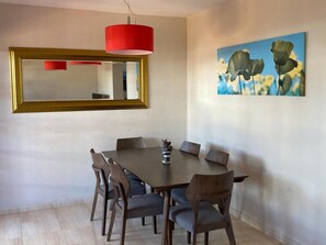 Un coin repas avec une table ronde, six chaises, un luminaire suspendu rouge, un grand miroir et un tableau représentant des fleurs accroché au mur.