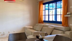 A living room with a beige sofa, a dining table with chairs, a window with blue shutters and orange curtains, and an air conditioning unit. - Villa Maria Luisa near the beach with private pool and Wi-Fi (Playa Blanca)