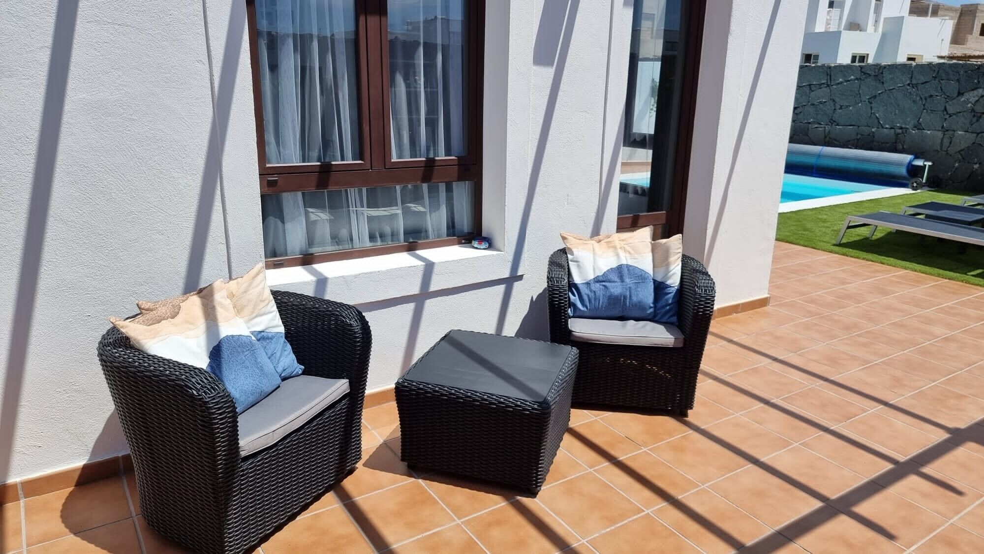 A patio with two wicker chairs and a small table, a window with curtains, and a pool in the background.
