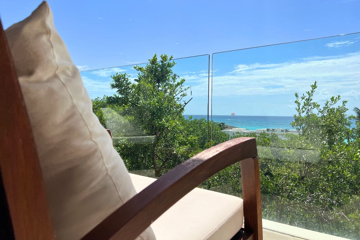 A wooden chair with a cushion, overlooking a balcony with a clear view of the ocean and a red umbrella in the distance.