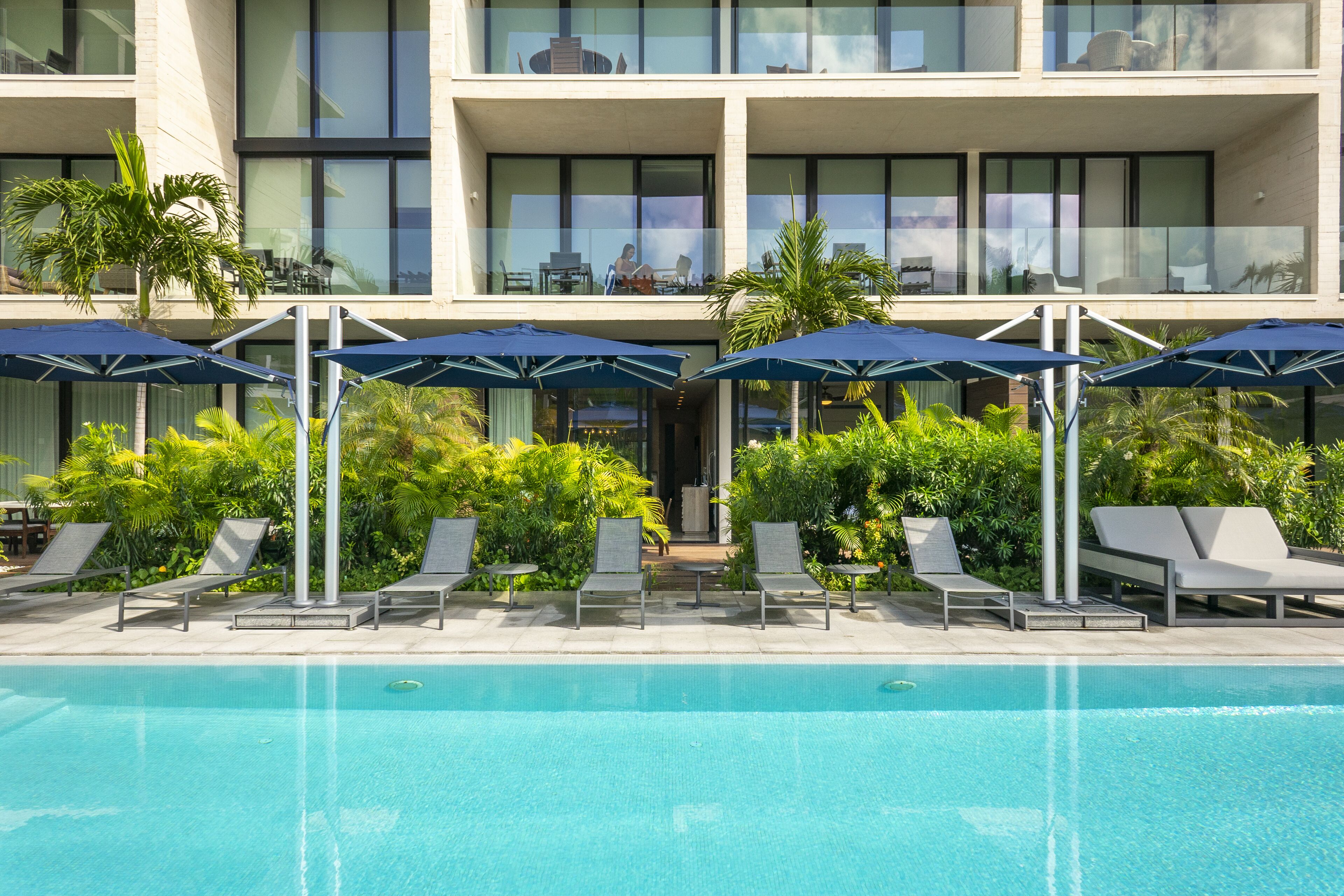 A pool area with lounge chairs and umbrellas, a building with large windows, and a well-maintained garden.