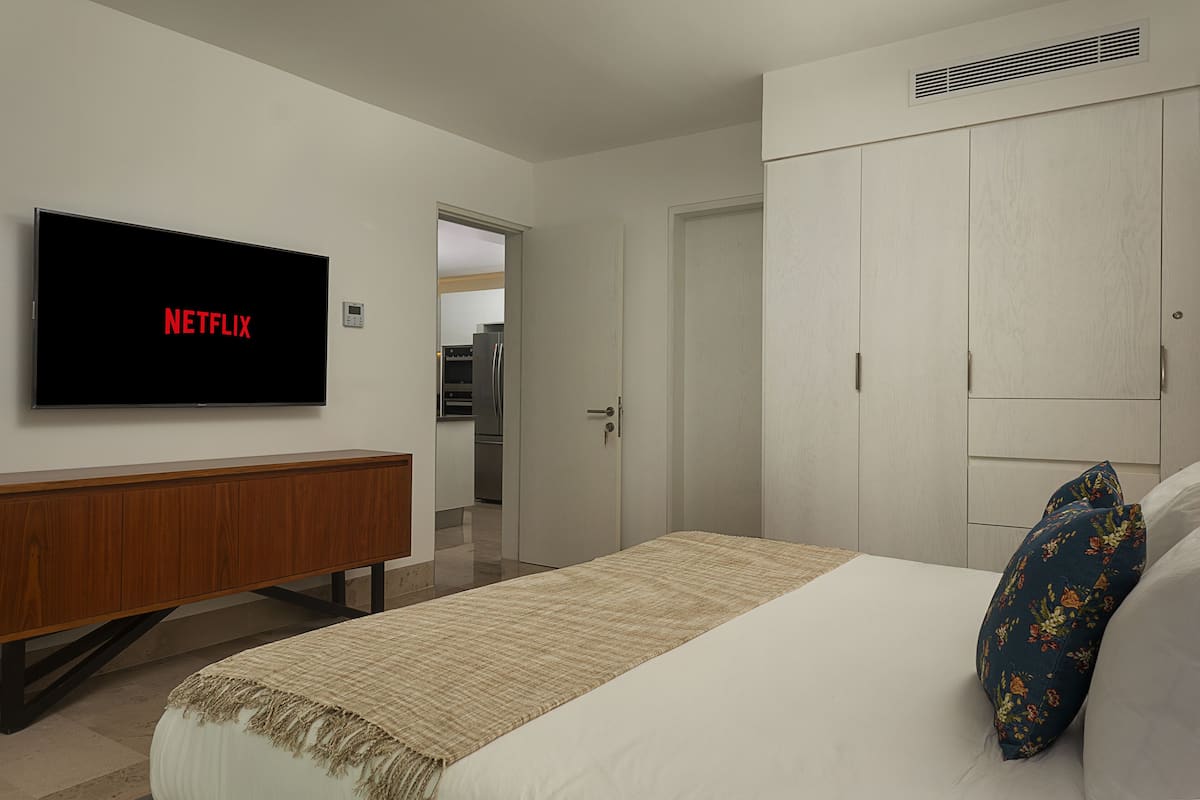 A bedroom with a bed, a TV mounted on the wall, a wardrobe, and a door leading to a kitchen.