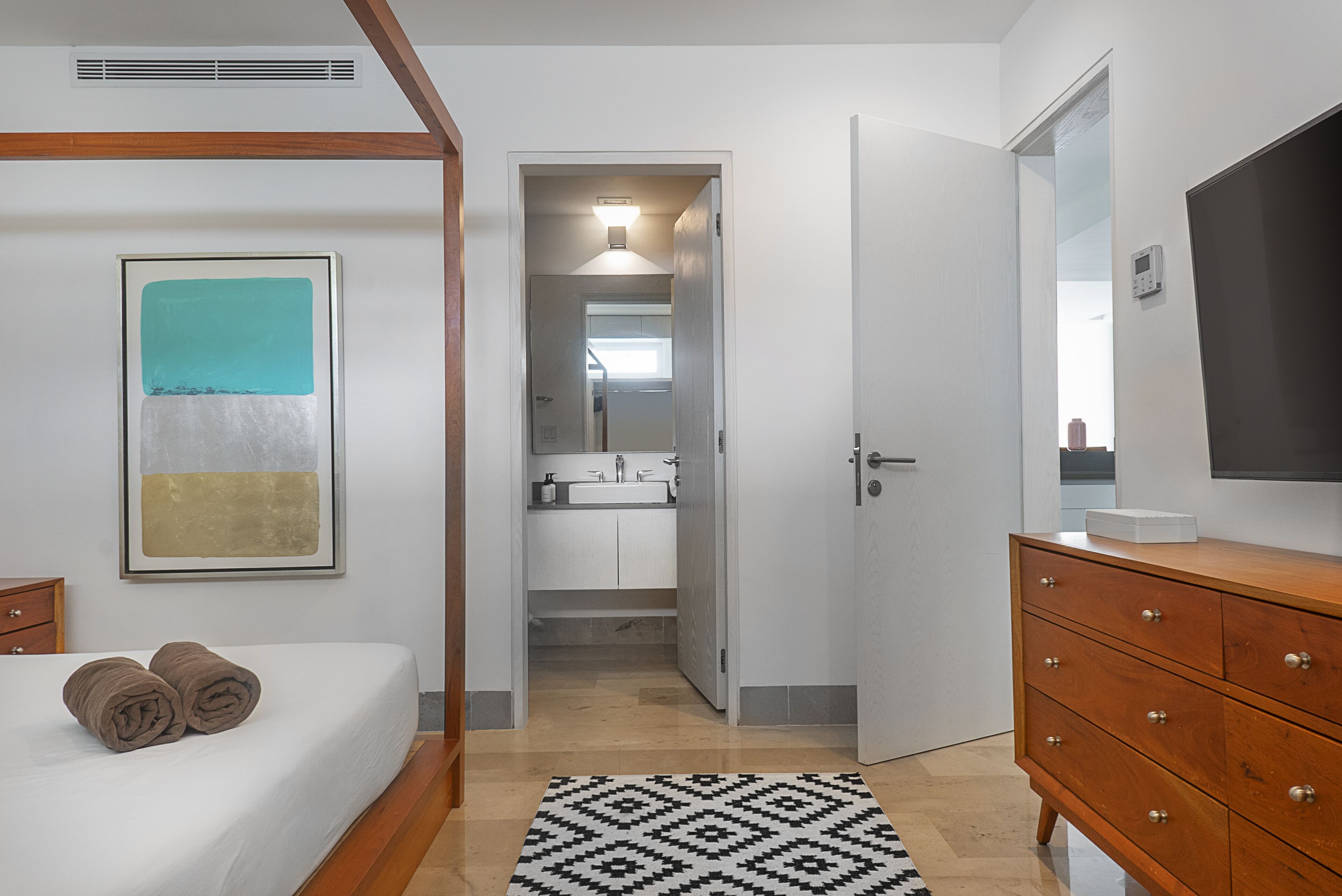 Un dormitorio con cama, cómoda de madera, baño visible a través de una puerta abierta y un cuadro abstracto enmarcado en la pared.