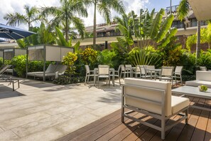Un patio al aire libre con sillones y mesas, rodeado de plantas tropicales y un edificio al fondo.