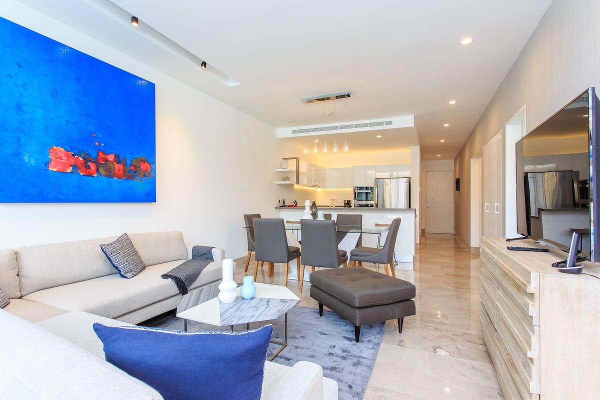 A modern living room with a sofa, ottoman, and coffee table, a dining area with chairs and a table, and a kitchen in the background.