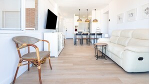 A modern living room with a sofa, a wicker chair, a dining area with chairs and a table, and a kitchen in the background.
