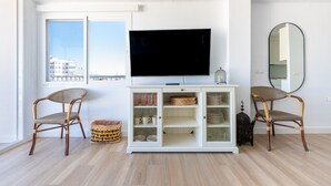 A modern living room with a large flat-screen TV, wicker chairs, a white cabinet with glass doors, and a view of the city through large windows.