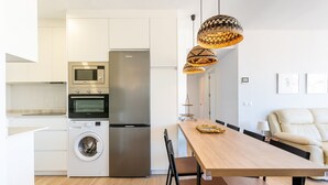 A modern kitchen with a stainless steel refrigerator, oven, microwave, and washing machine. There is a wooden dining table with chairs and a sofa in the adjacent living area.