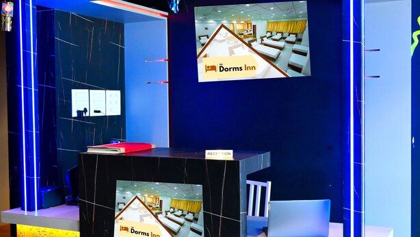 A reception desk with a digital screen displaying the hotel name, a laptop, and a white chair. - The Dorm Inn (Nashik)