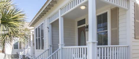 A white wooden house with a porch, a wooden deck, and a palm tree.