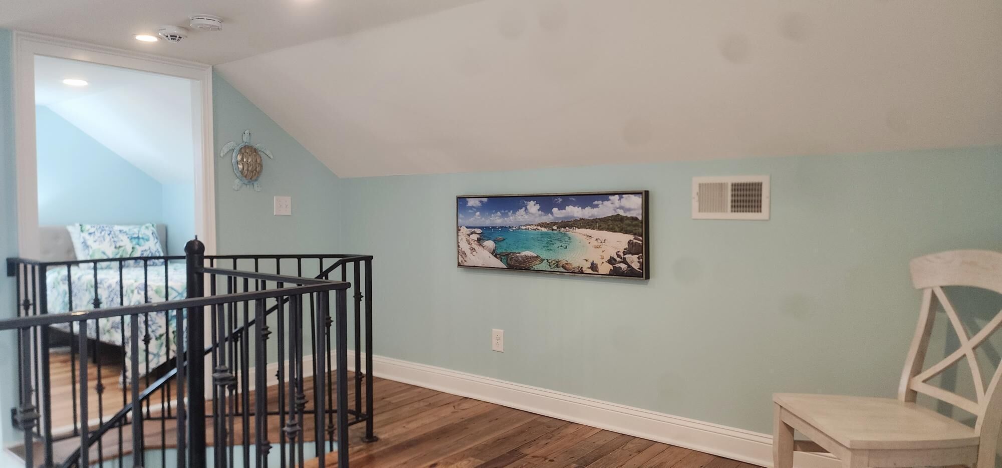 A room with a wooden floor, a black railing, a framed beach painting, and a white chair.