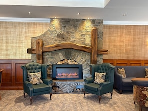A stone fireplace with a fire, flanked by two green armchairs with patterned cushions, and a dark sofa with a floral-patterned ottoman. - Burrendale Hotel Country Club & Spa (Newcastle)