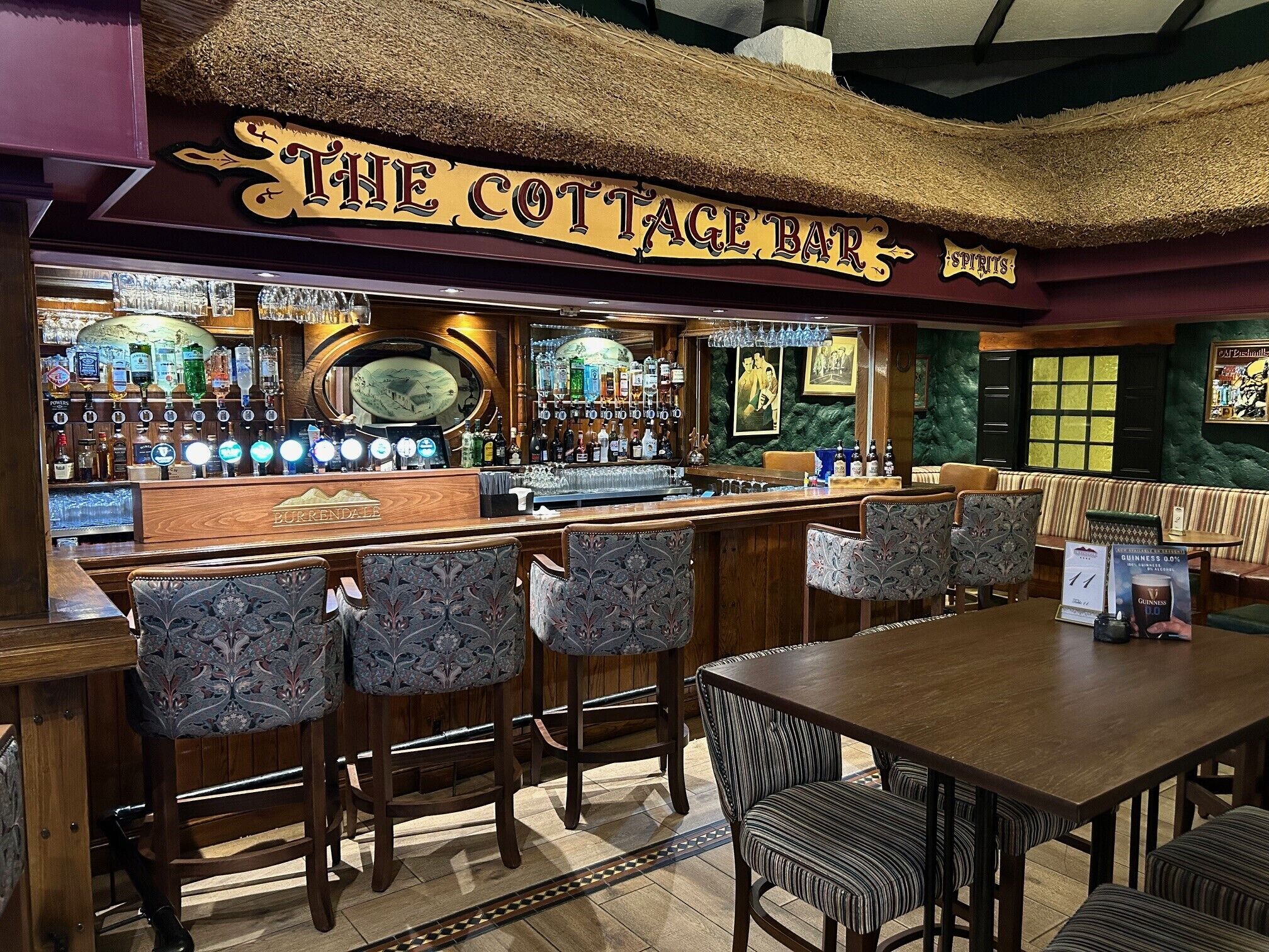 A bar area with a sign reading 'The Cottage Bar,' featuring a bar counter, multiple stools, and a table with chairs.