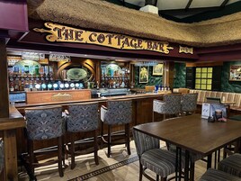 Zona de bar con un cartel que dice 'The Cottage Bar', que incluye una barra, varios taburetes y una mesa con sillas.
