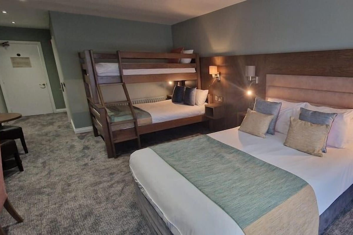 A hotel room with a bunk bed, a single bed, two bedside tables with lamps, a carpeted floor, and a door in the background.