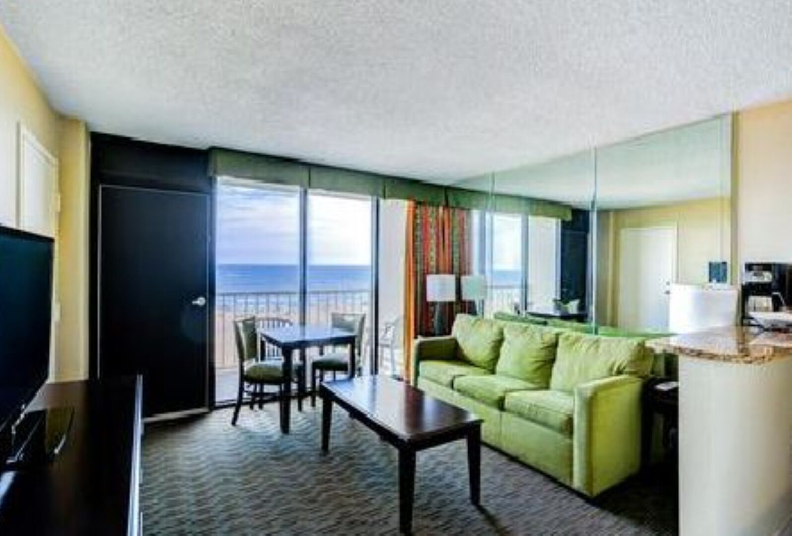 A hotel room with a balcony, a green sofa, a dining table, and a TV.