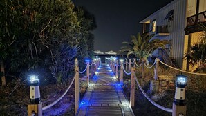 A wooden pathway with blue lighting, flanked by rope barriers and illuminated posts, leading to a building with a balcony. - Stunning Waterfront Home w/ Private Saltwater Pool & beautiful Tiki Bar! (Pensacola Beach)