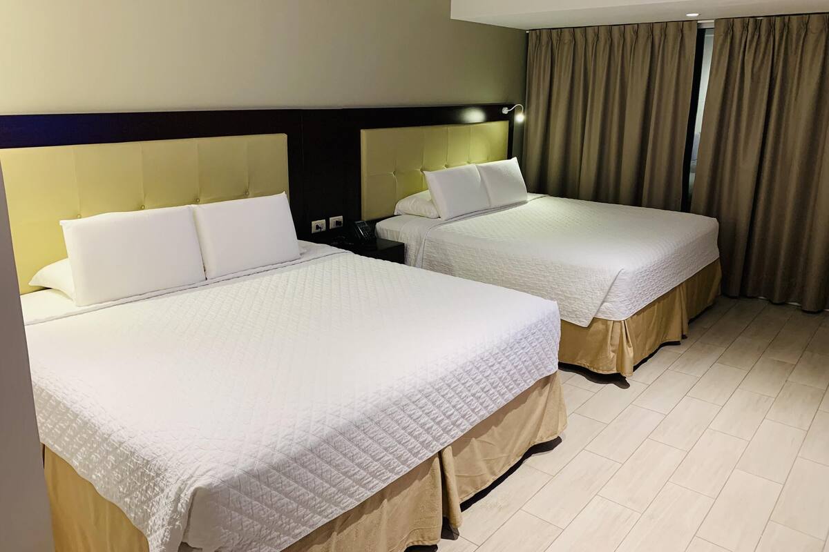 Habitación de hotel con dos camas, cortinas color beige y piso de baldosas claras.