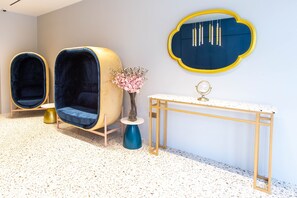 Een kamer met twee blauwe fauteuils, een bijzettafeltje met een bloevas, een spiegel met gouden lijst en een witte console tafel met gouden lijst.