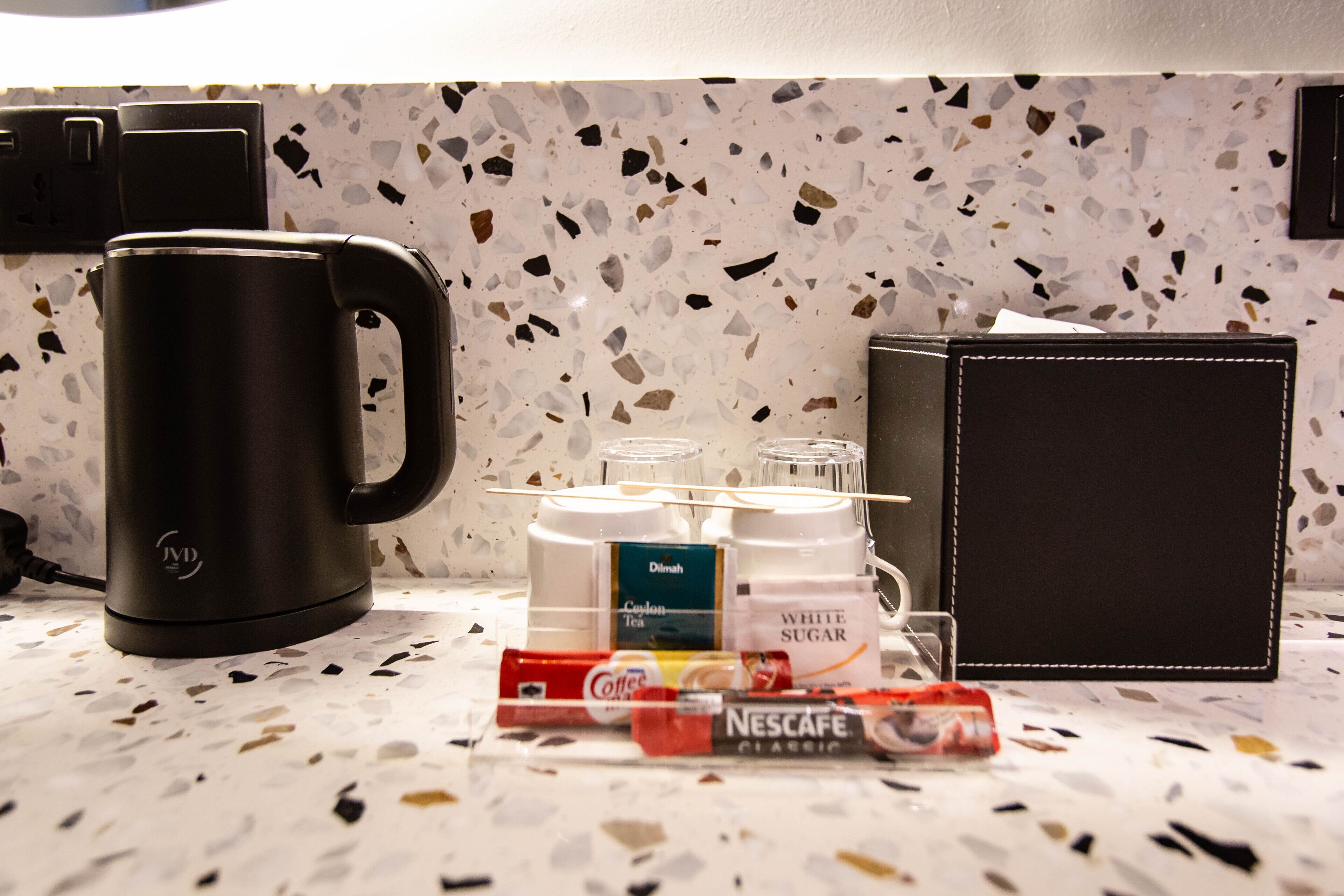 Una encimera de cocina con una cafetera, una caja de café Nescafe Classic, una caja de té Delish, un recipiente con azúcar blanca y un vaso de agua.