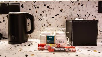 Una encimera de cocina con una cafetera, una caja de café Nescafe Classic, una caja de té Delish, un recipiente con azúcar blanca y un vaso de agua.