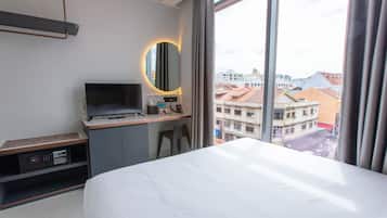 Habitación de hotel con una cama, un escritorio con televisor, un espejo y vistas a la ciudad a través de amplias ventanas.