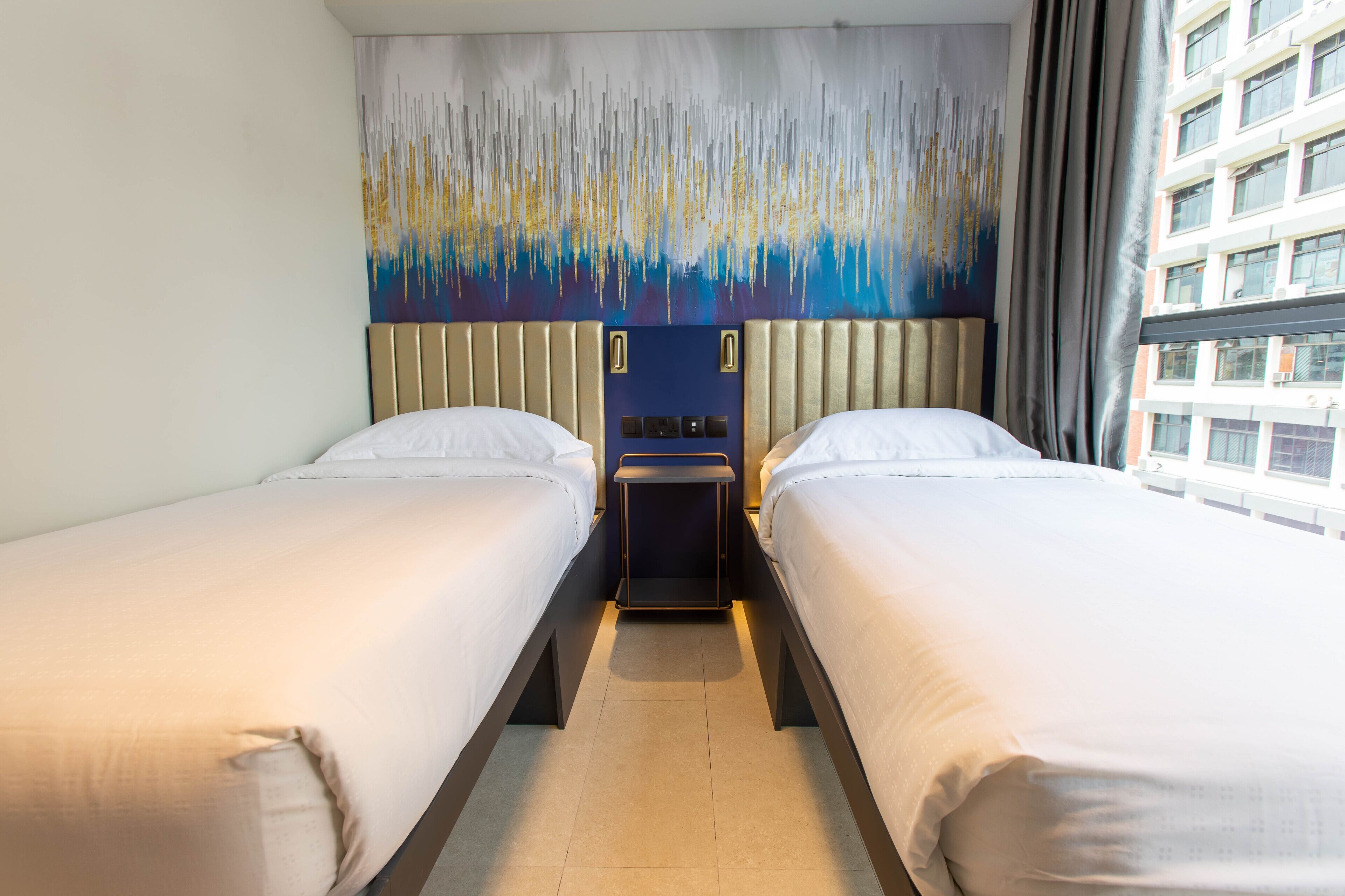 A hotel room with two beds, a headboard with a vertical light strip design, a small bedside table, and a window with a view of a building.
