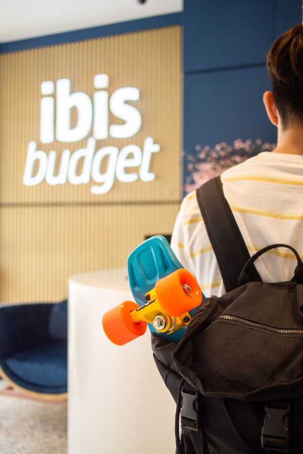 Eine Person mit Rucksack und Spielzeug-Skateboard vor einem Schild mit der Aufschrift 'ibis budget'.