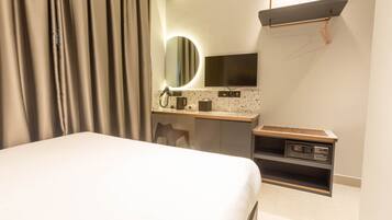 Quarto de hotel com cama, escrivaninha com espelho, televisão e prateleira pequena.
