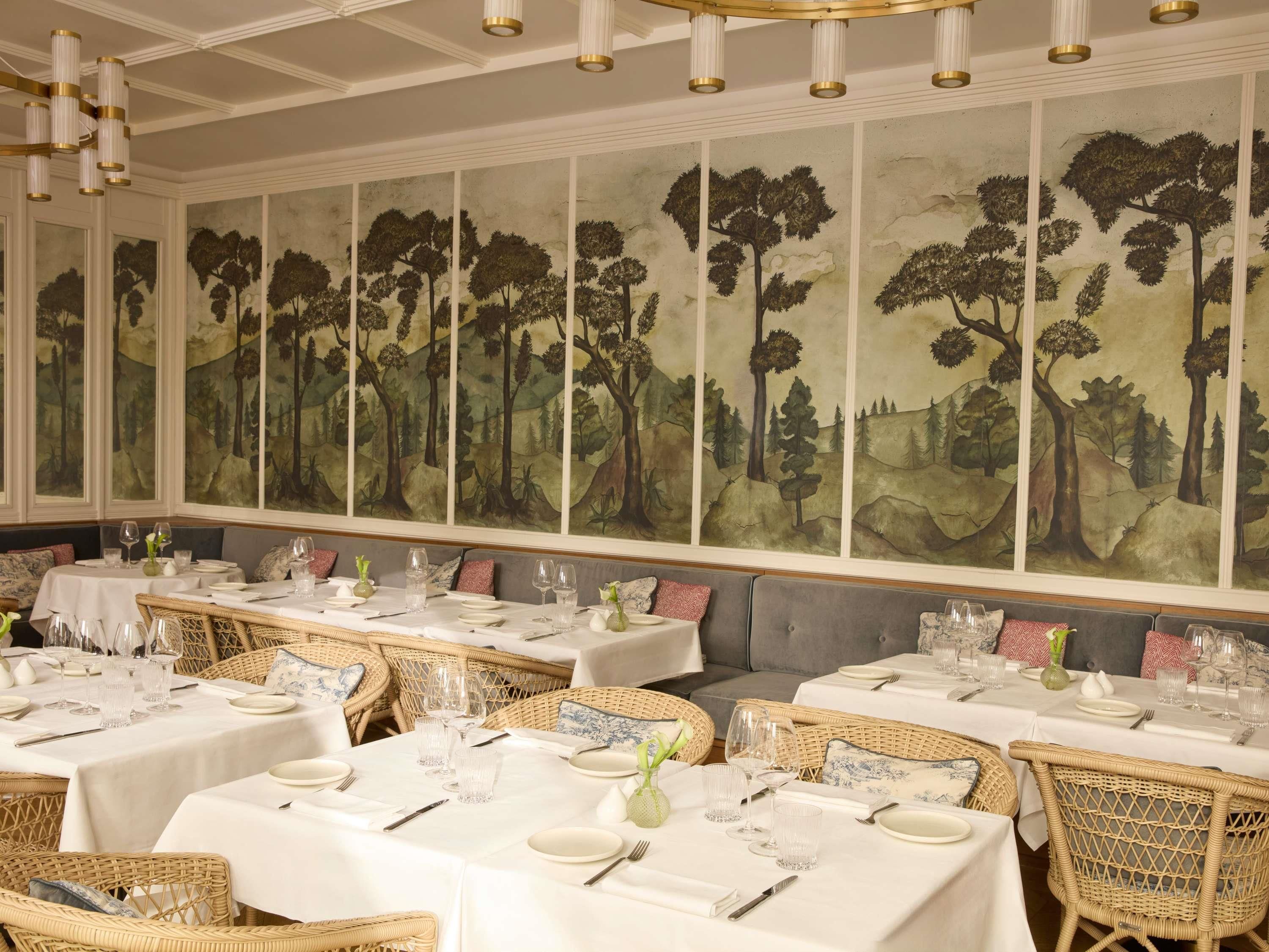 Un espace repas avec des tables dressées pour le service, une grande fresque représentant une forêt sur le mur et un lustre.