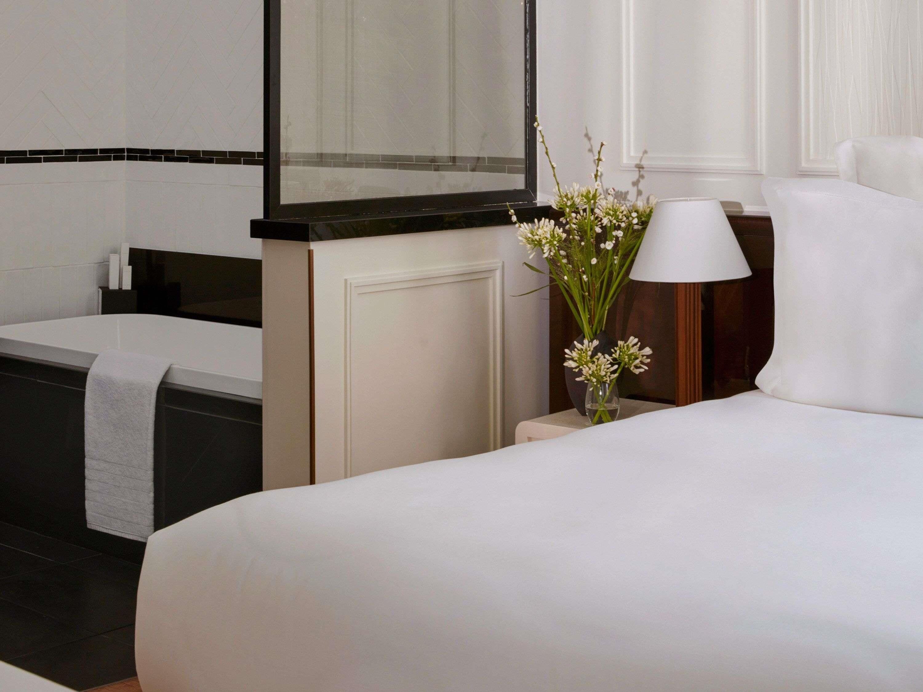 Habitación de hotel con cama, mesita de noche con lámpara, un jarrón de flores y una bañera.
