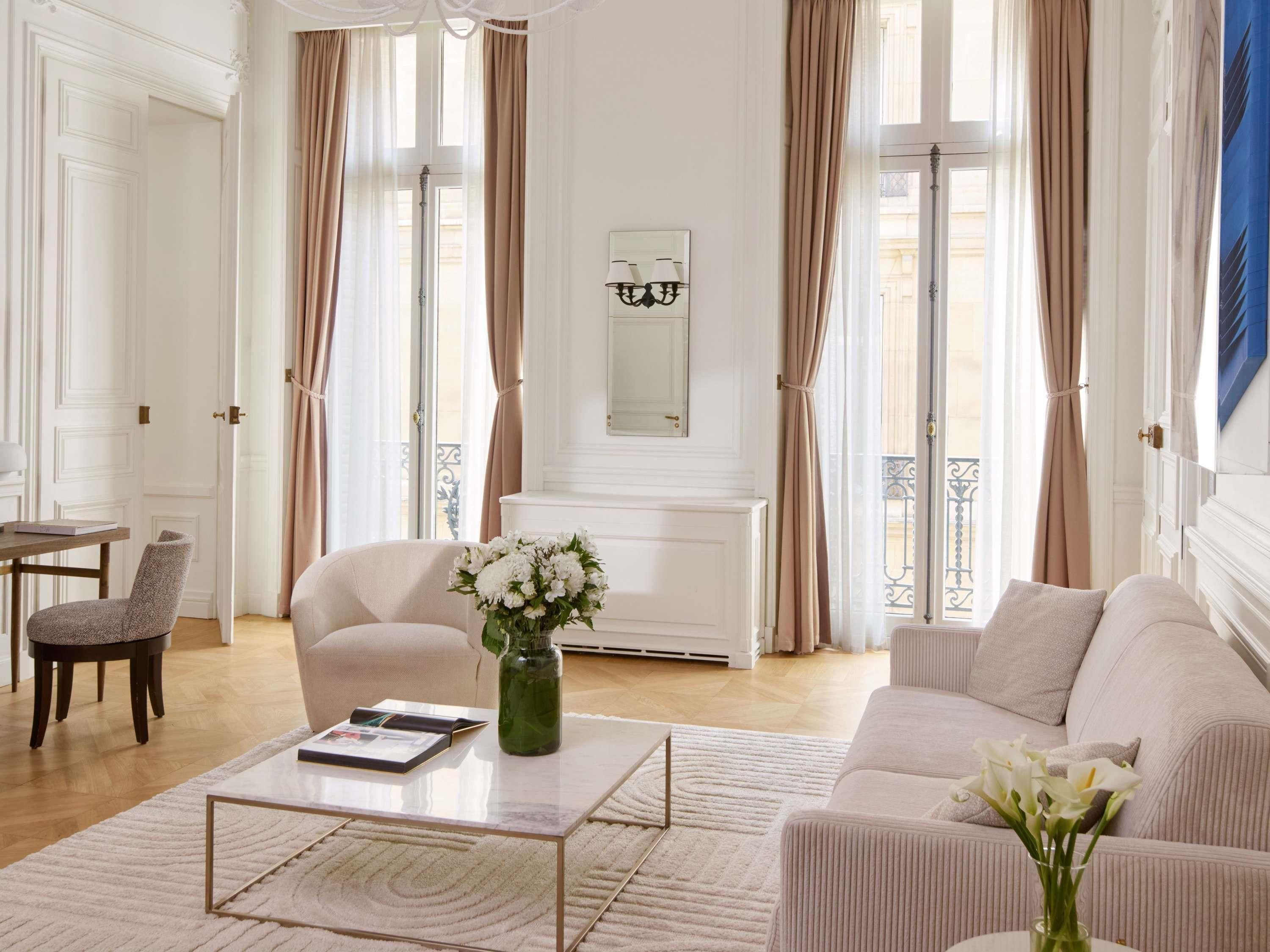 A spacious living room with a sofa, armchair, and coffee table, large windows with curtains, and a radiator.
