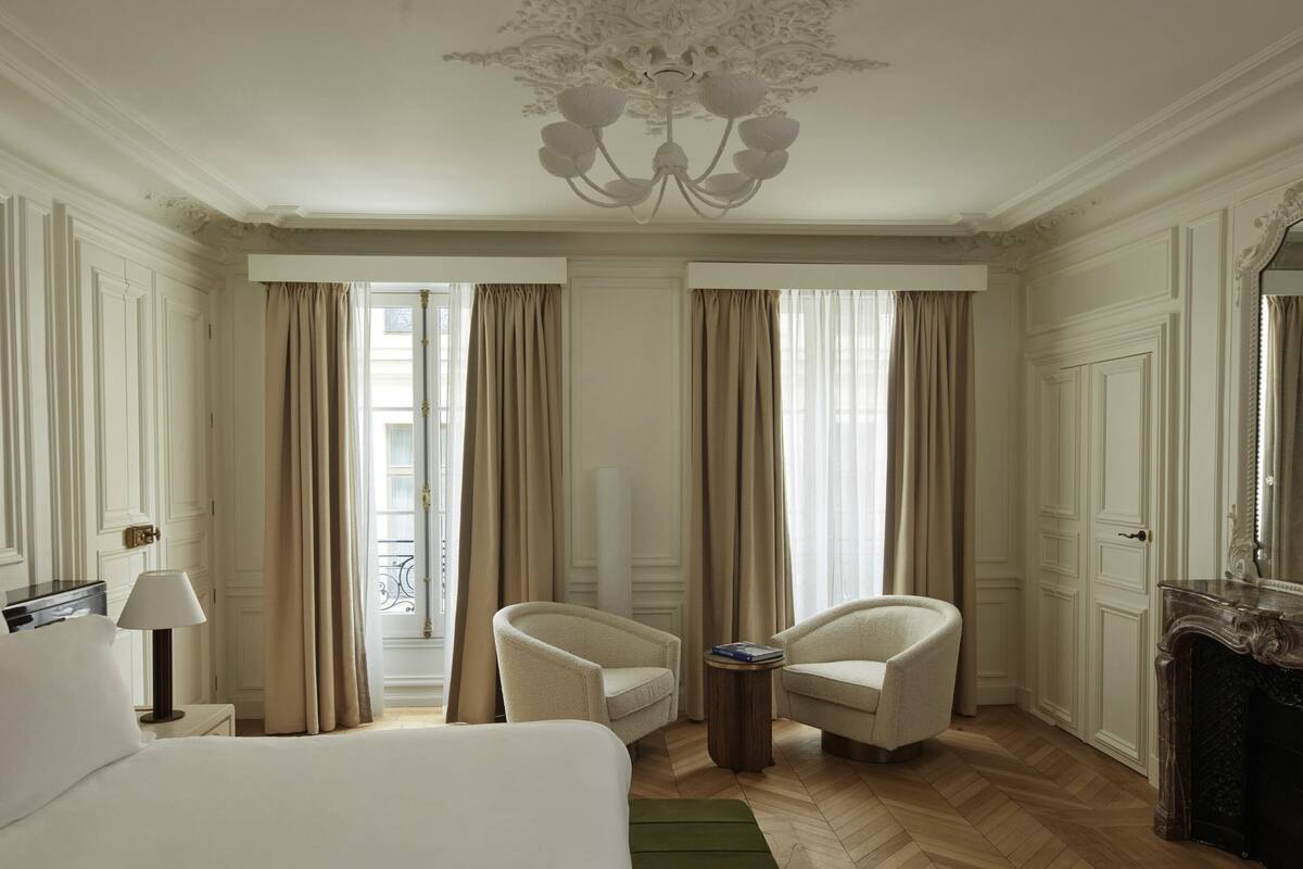 A hotel room with a large bed, two armchairs, a small side table, a green rug, a chandelier, and a fireplace.