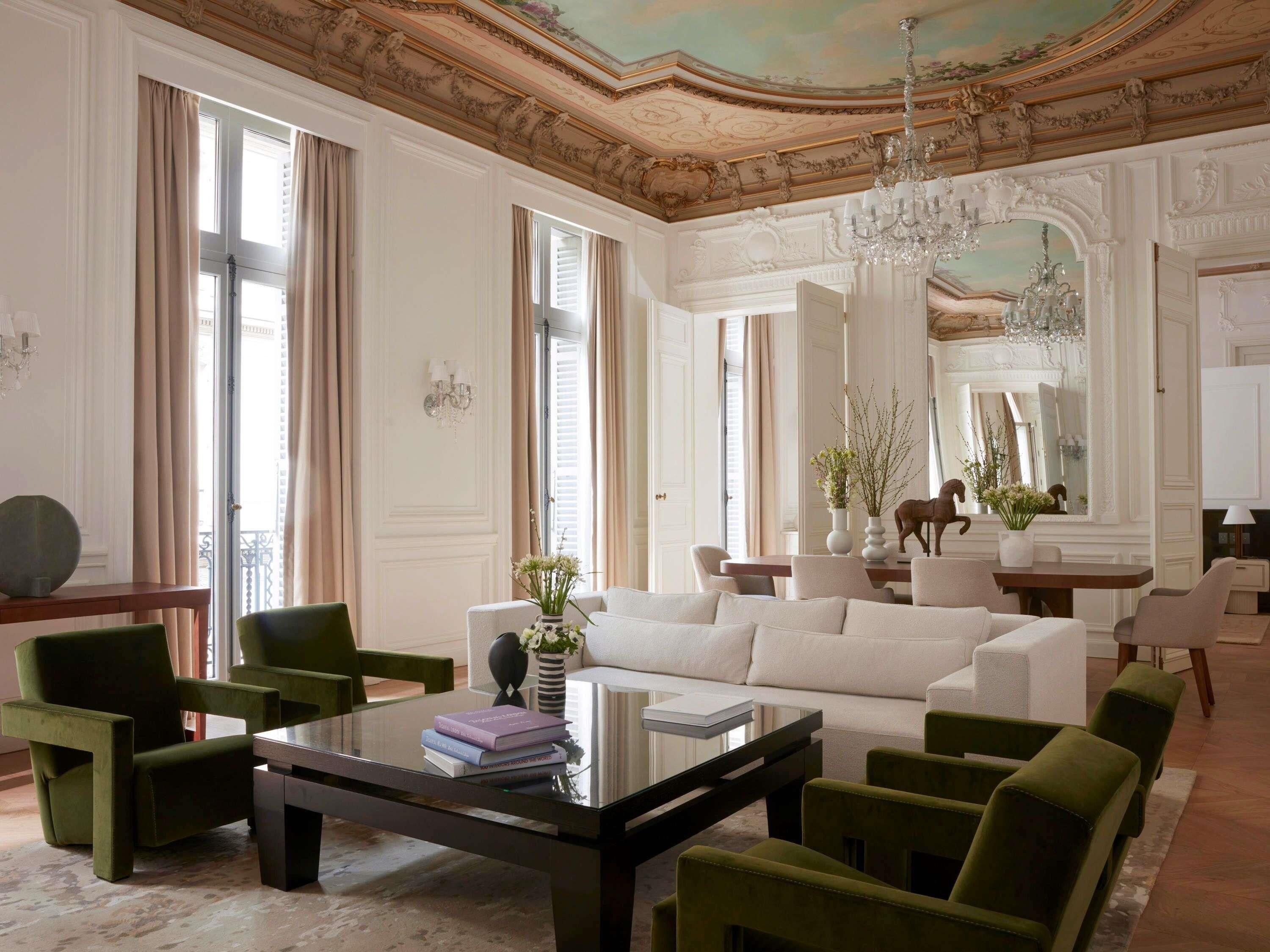 A spacious living room with a large white sofa, green armchairs, a glass coffee table, and a chandelier.