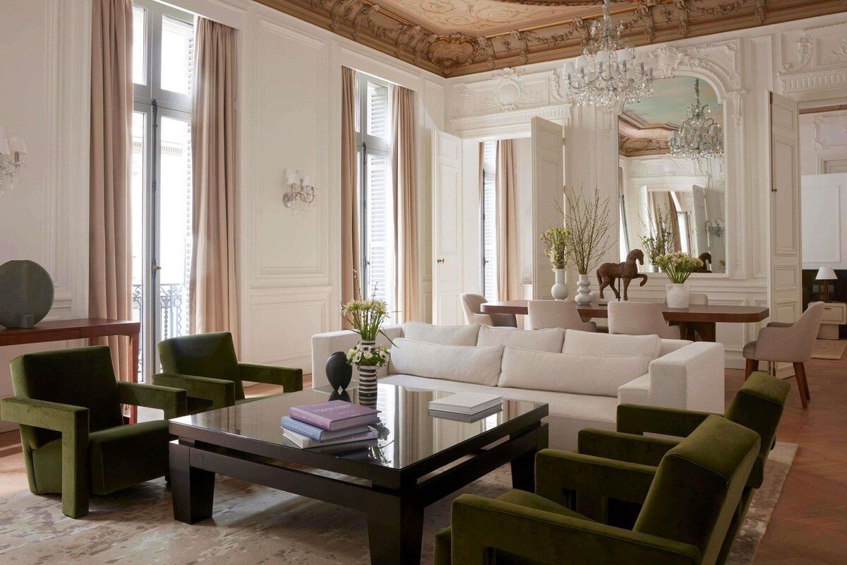 A spacious living room with a large white sofa, green armchairs, a glass coffee table, and a chandelier.