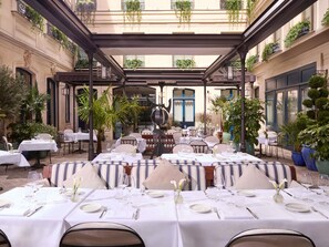 Un espacio de comedor al aire libre con mesas preparadas para dos, rodeado de follaje y detalles arquitectónicos.