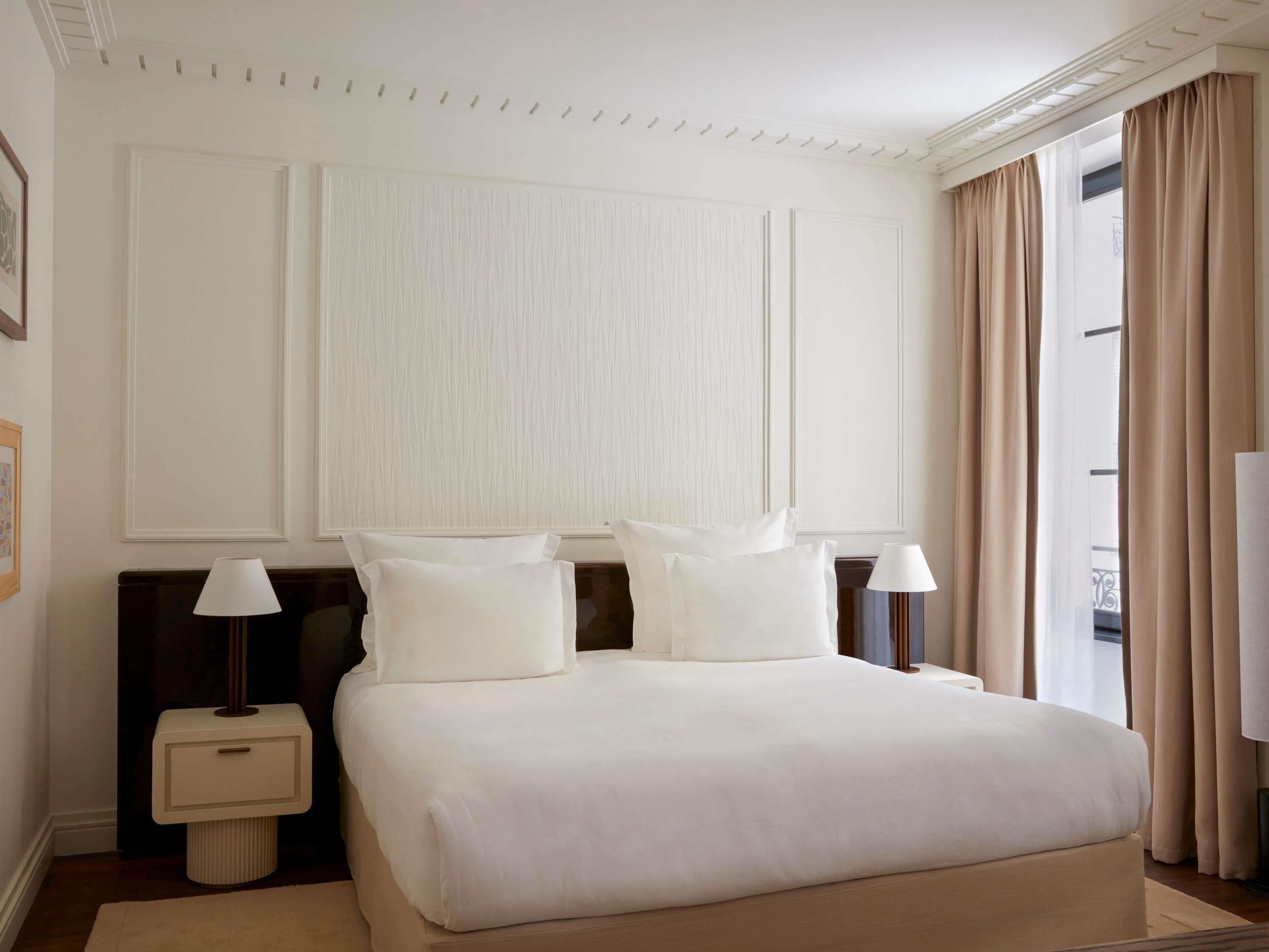 A hotel room with a large bed, two bedside tables with lamps, a large window with curtains, and a wall with paneling.