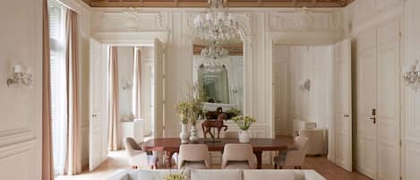 A spacious room with a high ceiling, ornate moldings, a chandelier, a dining area with a table and chairs, and a sofa with vases.