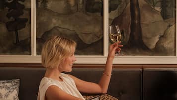 A person seated on a wicker chair, holding a glass of white wine, with a painting of a forest behind them.